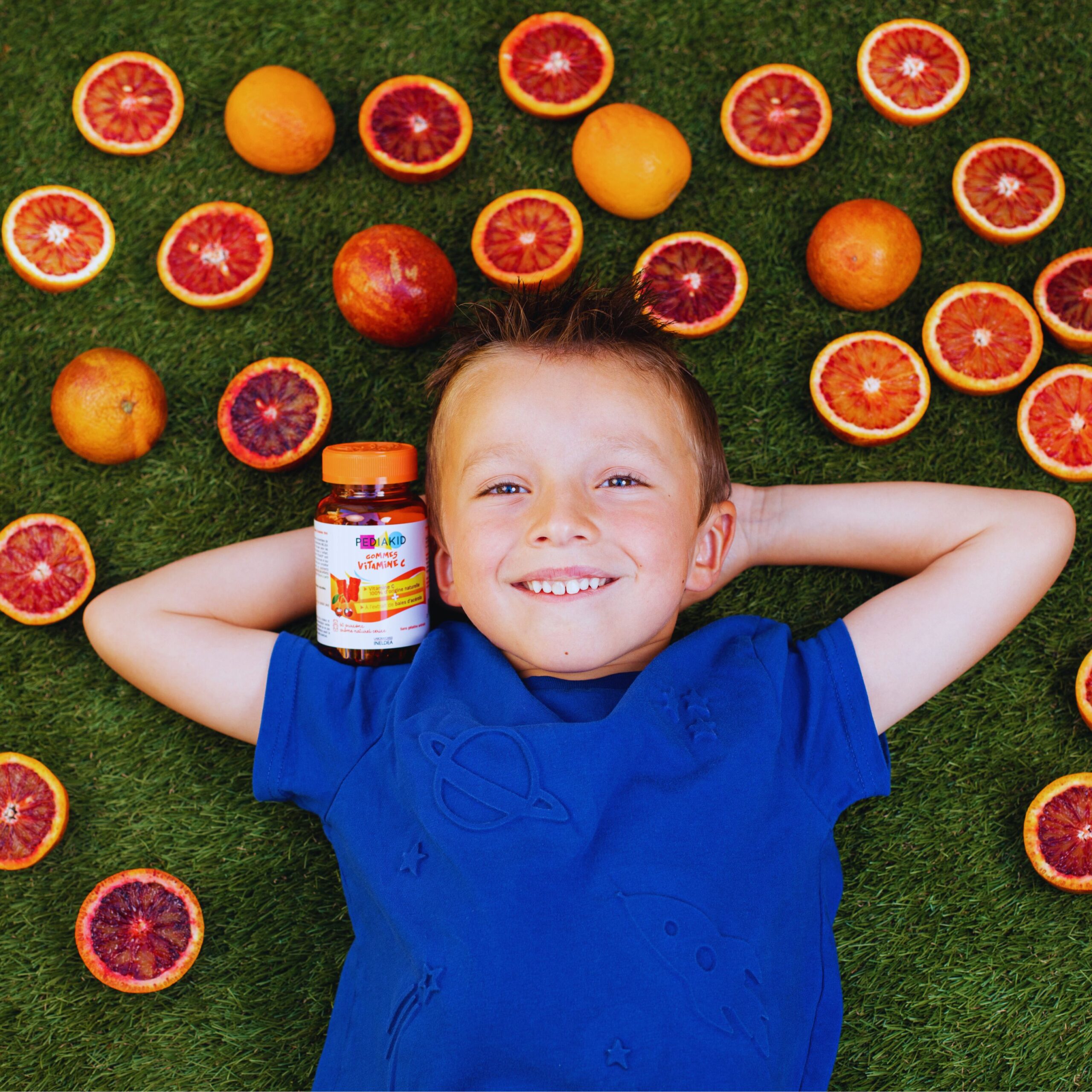 Glückliches Kind liegt auf dem Gras mit PEDIAKID Vitamin C, umgeben von frischen Blutorangen – natürliche Unterstützung für das Immunsystem